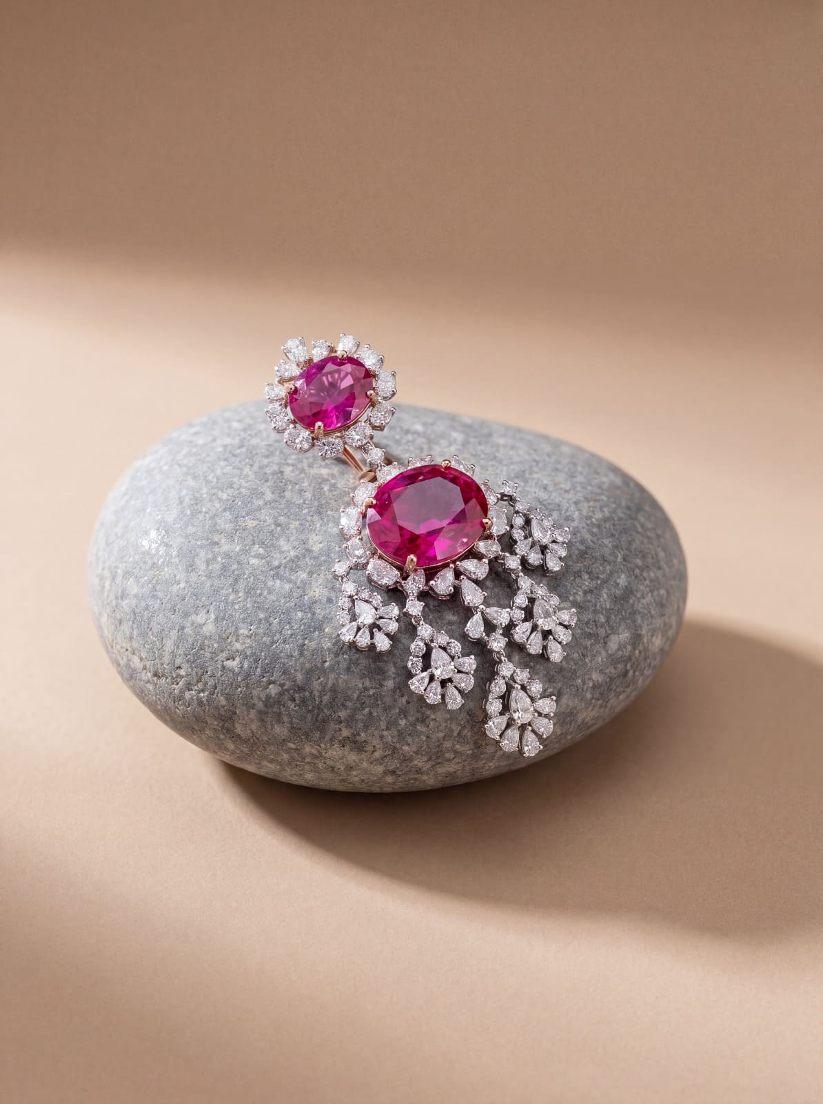 AI jewellery packshot: a ruby and diamond piece, a stone-set flower above a cascading drop, resting on a smooth grey stone against a sand-coloured background.
